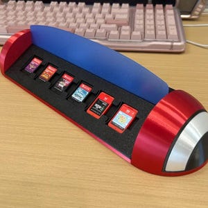 May include: A red, blue, and white plastic game cartridge storage stand shaped like a Poké Ball. The stand features a black foam insert with six slots, each holding a small rectangular game cartridge. The design includes a curved blue back panel.