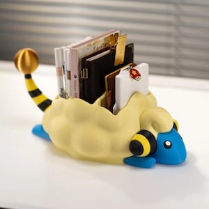 May include: A desk organiser shaped like a blue and yellow creature with a fluffy body, featuring a tail with a gold sphere, holding various notebooks, pens, and a white card with a small red mushroom charm attached to the top edge of the card.