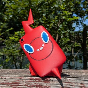 May include: A bright red, rectangular-shaped figure with a lightning bolt-shaped protrusion on top and a pointed base. The figure features two large, oval-shaped blue eyes, a circular black outline, and a small white mouth with two visible teeth.