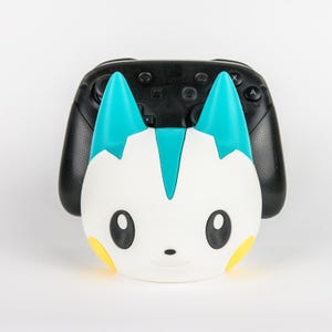 Könnte beinhalten: Ein weißer, türkisfarbener und gelber Controller-Ständer in Charakterform mit großen dunklen Augen und spitzen Ohren. Der Ständer steht vor einem schwarzen Nintendo Switch Pro Controller, der einen strukturierten Griff und Tasten hat.
