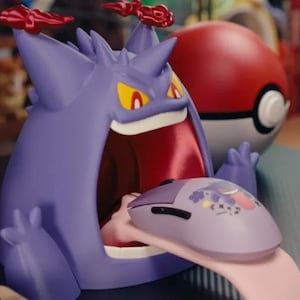 May include: A purple Gengar figure with red accents on its head and yellow eyes, featuring a wide-open mouth. A light purple computer mouse with a Gengar graphic rests on the figure's long pink tongue, positioned in front of a red and white Poké Ball.