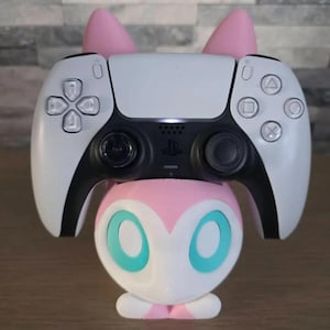 May include: A white and black game controller rests on a pink and white character-shaped stand. The stand features large teal oval eyes and two pointed pink ears extending upward behind the controller, creating a decorative display.
