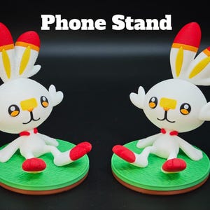 Puede incluir: Dos figuras blancas parecidas a conejos con puntas de orejas rojas y amarillas, parches nasales naranjas y ojos negros, cada una sentada sobre una base circular verde. El texto Phone Stand está centrado sobre las figuras.