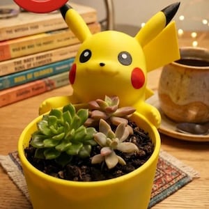 May include: A yellow planter shaped like the character Pikachu, featuring pointed ears with black tips, red circular cheeks, and black eyes. The planter holds two small green and light purple succulent plants in dark soil.