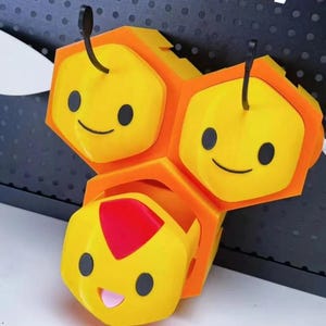 Pode incluir: Uma figura amarela de três partes em forma de favo de mel com olhos pretos e bocas sorridentes. As duas seções superiores possuem antenas pretas, enquanto a seção inferior apresenta uma marca triangular vermelha e uma língua rosa.