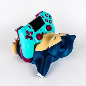 Könnte beinhalten: Ein türkisfarbener und dunkelvioletter Videospiel-Controller, der auf einer dunkelblauen und cremefarbenen Snorlax-Figur liegt. Der Controller verfügt über ein schwarzes zentrales Touchpad, dunkelviolette Analogsticks und violette Richtungstasten.