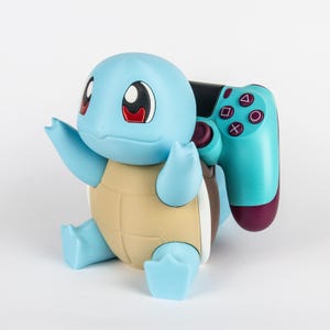 May include: A light blue and tan figurine of a turtle-like creature with large eyes, holding a teal and black video game controller. The controller features standard circular action buttons with purple symbols and a black directional pad.
