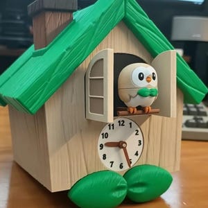 May include: A cuckoo clock shaped like a house with a bright green roof, light tan walls, and two green leaves at the base. A small, round, tan and white owl figure with a green bowtie stands in the open window above a white clock face with black numbers.