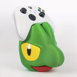 Puede incluir: Un mando de videojuego inalámbrico blanco sobre un soporte con forma de personaje en colores verde, amarillo y rojo. El soporte presenta una cabeza verde estilizada con grandes ojos amarillos, una pupila negra y una boca roja para el mando.