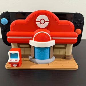Può includere: Una docking station per console da gioco a forma di edificio rosso, bianco e blu con un emblema centrale a forma di Poké Ball. Il supporto presenta una base color sabbia, una porta blu e un piccolo cabinato arcade rosso sul lato sinistro.