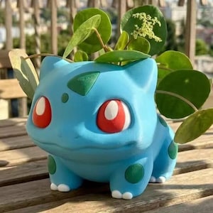 Könnte beinhalten: Ein hellblauer, Bulbasaur-förmiger Übertopf mit dunkelgrünen Flecken, roten Augen mit weißen Pupillen und weißen Krallen. Eine grüne Pilea peperomioides Pflanze mit runden Blättern und einer kleinen weißen Blütenrispe wächst aus der Figur heraus.