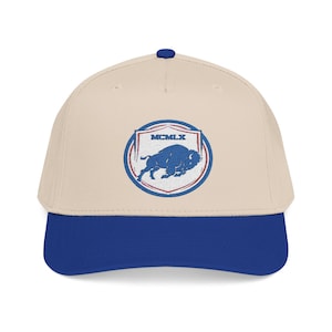 May include: A beige baseball cap with a royal blue brim and a top button. The front features an embroidered circular patch with a blue buffalo silhouette, red and blue border lines, and the Roman numeral text MCMLX centered at the top.
