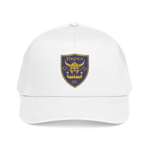May include: A white baseball cap featuring a centered shield-shaped emblem. The emblem has a dark blue background with a gold-colored Viking helmet, two crossed axes, and a longship graphic. The word VIKINGS is printed in gold above the helmet.