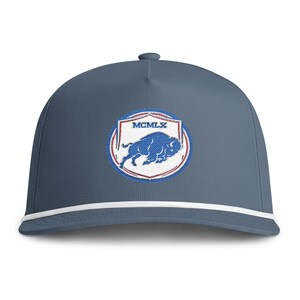 May include: A slate blue baseball cap featuring a white rope detail across the base of the crown. The front displays an embroidered circular patch with a red and blue border, a blue buffalo graphic, and the Roman numeral text MCMLX in white.