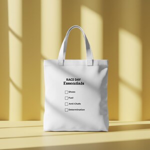 May include: A white tote bag featuring the black printed text RACE DAY Essentials followed by a checklist with four empty square boxes next to the words Shoes, Fuel, Anti-Chafe, and Determination. The bag has two matching white fabric handles.
