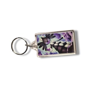 May include: A rectangular keychain featuring a graphic illustration of a dark-colored creature with white accents and purple highlights. The character has a spiky head and a long, segmented body, set against a swirling purple and blue background.