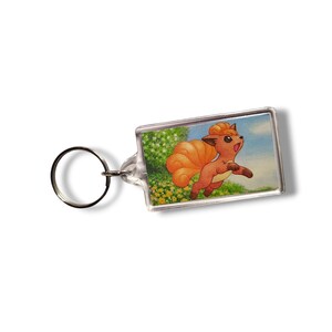 May include: A rectangular clear acrylic keychain featuring an illustration of a small, orange, fox-like creature with a large, curly tail. The character is depicted running through a field of yellow flowers under a bright blue sky.