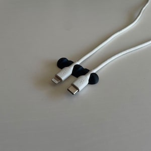 Op de afbeelding: Een zwarte siliconen kabelorganizer met drie afgeronde sleuven die twee witte oplaadkabels vasthouden. De ene kabel heeft een Lightning-connector en de andere een USB-C-connector, beide met het Belkin-logo in reliëf op de stekkerkoppen.