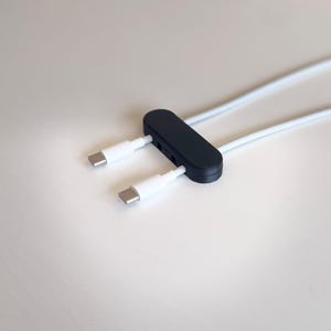 May include: A black cable management organiser with an elongated oval shape, featuring two white USB-C charging cables threaded through the device. The organiser holds the cables in place, with the metallic USB-C connectors extending outward.