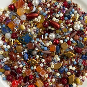 May include: A large collection of assorted loose beads in various shapes, sizes, and textures. The mix includes glass, stone, and plastic beads in shades of red, orange, blue, brown, white, and gold, featuring faceted, round, and geometric forms.