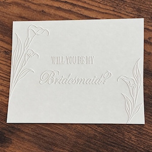 May include: A rectangular white card featuring an embossed design of calla lilies on the left and right sides. The center of the card displays the embossed text "WILL YOU BE MY Bridesmaid?" in a combination of block and script lettering styles.