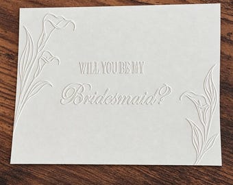 May include: A rectangular white card featuring an embossed design of calla lilies on the left and right sides. The center of the card displays the embossed text "WILL YOU BE MY Bridesmaid?" in a combination of block and script lettering styles.