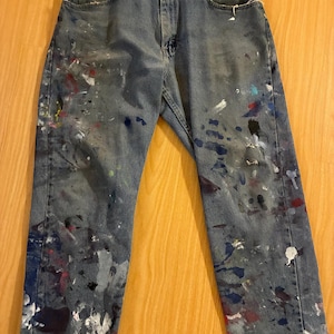 May include: A pair of medium-wash blue denim jeans featuring a button fly closure and five pockets. The fabric is heavily splattered with various paint colours including white, red, blue, and black, covering the front legs from the thighs down.