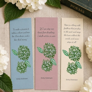 May include: Three rectangular paper bookmarks in light blue, dusty rose, and cream. Each features a watercolor illustration of green hydrangea flowers and a quote by Emily Dickinson. The text includes the author's name at the bottom of each bookmark.