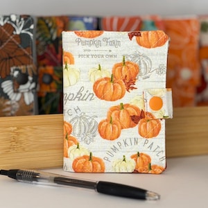 May include: A fabric-covered notebook featuring a white background with orange pumpkin illustrations, green leaves, and the text Pumpkin Farm Pick Your Own Pumpkin Patch. The cover includes a small orange snap button closure and a black ink pen.