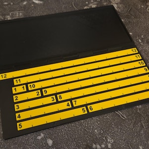 May include: A black rectangular board featuring a series of horizontal yellow strips arranged in a staggered pattern. Each yellow strip is marked with black numbers ranging from 1 to 12, indicating a sequence or measurement scale on the surface.