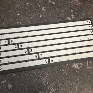 Op de afbeelding: Een rechthoekig zwart spelbord met meerdere horizontale witte banen. Elke baan is gelabeld met numerieke reeksen van 1 tot 12, gerangschikt in rijen van variërende lengtes en gemarkeerd met kleine verticale streepjes.