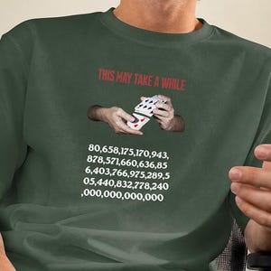 May include: A dark olive green t-shirt featuring the red text "THIS MAY TAKE A WHILE" above a graphic of hands shuffling a deck of playing cards, followed by a long sequence of white numbers representing a large mathematical probability.