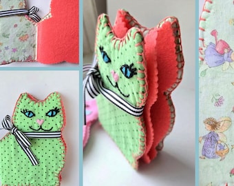 May include: A handmade cat-shaped needlebook with a green polka dot exterior and a pink interior. The needlebook is adorned with a black and white striped ribbon and features embroidered details. The text "Kitty NeedleBook" is visible.