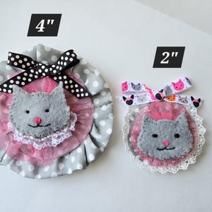 May include: Two felt cat brooches with pink tulle and gray felt. The larger brooch is 4 inches in diameter and the smaller brooch is 2 inches in diameter.