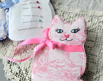 May include: Handmade cat-shaped pin cushions. One is white with pins, the other is pink with a pink ribbon and a vintage illustration. The cat's face has blue eyes and a pink nose. The pin cushions are on a lace doily.