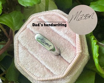 May include: A silver-toned metal ring featuring a flat, rectangular face engraved with the name Mason in cursive script. The ring is displayed in a pale pink, octagonal velvet ring box. A circular insert shows the original handwriting sample.