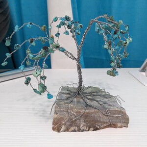 May include: A decorative wire tree sculpture featuring twisted silver-colored metal branches adorned with small, irregular turquoise-colored stone chips. The tree is anchored to a flat, rectangular, multi-toned brown and gray stone base.