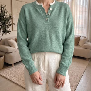 Può includere: Un maglione in maglia a maniche lunghe color verde salvia con scollo a henley dotato di tre bottoni chiari e bordi a coste su colletto, polsini e orlo. Il maglione è indossato sopra una maglietta bianca a girocollo e pantaloni bianchi.