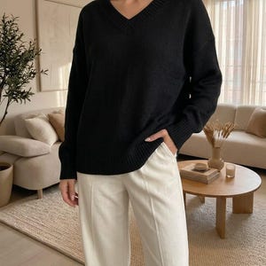 Op de afbeelding: Een zwarte trui met lange mouwen en een V-hals met geribbelde boorden bij de kraag, mouwen en zoom, gecombineerd met een off-white broek met rechte pijpen en een zichtbare verticale naad in het midden van elke broekspijp.