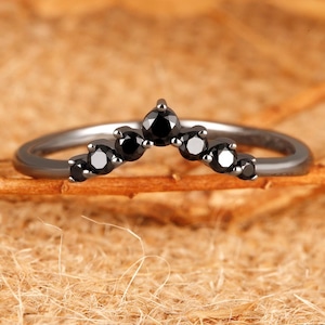 May include: A V-shaped ring crafted from dark metal, featuring a series of seven round-cut black stones set along the curved band. The stones are arranged in a graduated size pattern, with the largest stone positioned at the centre peak of the band.
