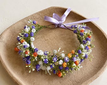 May include: A circular floral crown featuring small clusters of purple, white, and orange blossoms on a thin green wire frame. A light lavender satin ribbon is tied in a bow at the back, with two long ribbon tails extending from the floral arrangement.