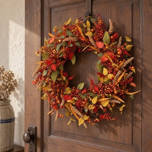 May include: A circular autumn wreath featuring a dense arrangement of red berries, orange and yellow leaves, and textured tan wheat-like stalks. The wreath is mounted on a dark brown wooden door with a visible metal door handle and hardware.
