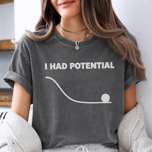 May include: A charcoal gray short-sleeve t-shirt featuring the white text I HAD POTENTIAL above a white graphic of a curved line and a circle representing a ball at the bottom of a slope, printed on the front of the garment.