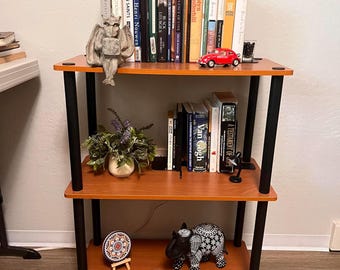 May include: A three-tier bookshelf with brown wood-grain shelves and black cylindrical supports. The shelves hold various books, a stone-colored gargoyle figurine, a red toy car, a potted plant, a small decorative plate, and a black and white patterned hippo figurine.