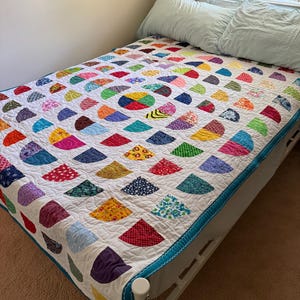 May include: A quilt featuring a white background with a repeating pattern of colorful, multi-patterned semi-circle shapes arranged in a circular design. The quilt has a solid teal blue border and is displayed on a bed with two light blue pillows.