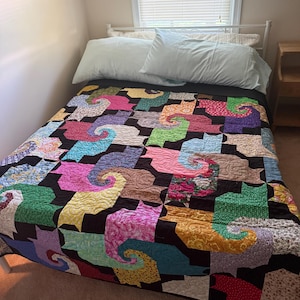 May include: A bed quilt featuring a repeating pattern of cat-shaped silhouettes in various colors and floral or geometric prints, separated by a black background. The quilt is spread over a bed with two light blue pillows and a white metal headboard.