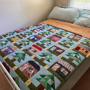 Puede incluir: Una colcha de patchwork con un patrón repetitivo de casas y árboles de hoja perenne hechos de retazos de tela en varios colores y estampados. La colcha tiene un fondo azul claro con bandas verdes y está doblada para mostrar el reverso naranja.