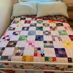 May include: A patchwork quilt featuring a grid of multi-colored fabric squares separated by white fabric with small black dots. The quilt is draped over a bed with a white metal frame and two light blue pillows positioned at the head of the bed.