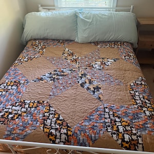 May include: A bed quilt featuring a star pattern composed of solid tan fabric panels and a patterned fabric displaying a repeating print of small cats in black, white, orange, and grey. The quilt is topped with two light blue pillows.