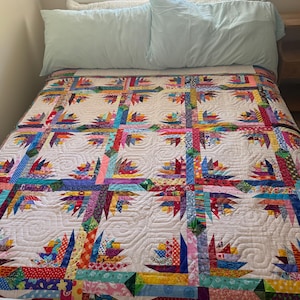 May include: A patchwork quilt featuring a repeating star-like pattern made from various colorful fabric scraps in shades of red, blue, yellow, green, and pink, set against a white background with intricate swirling machine-stitched quilting details.